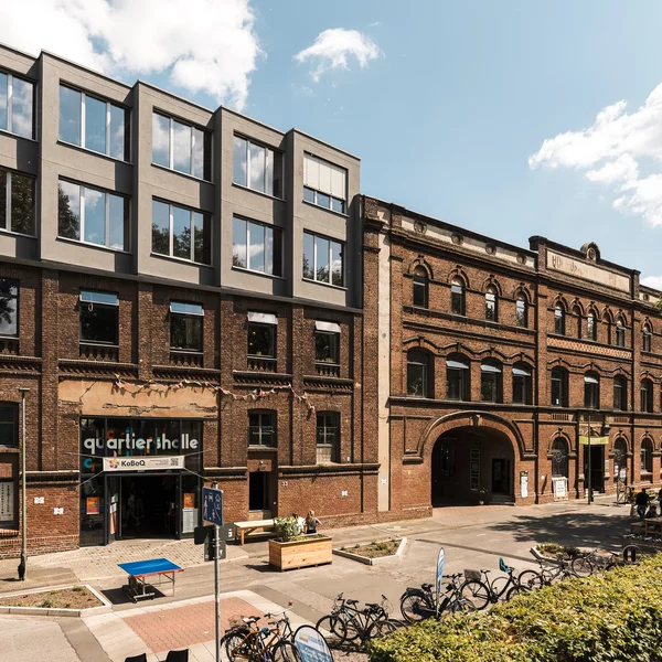 Außenansicht der KoFabrik in Bochum, ein Backsteingebäude mit einem zweigeschossigen grauen Aufbau auf der linken Gebäudehälfte. Die Stühmeyerstraße im Vordergrund ist verkehrsberuhigt.