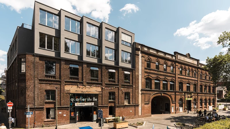 Außenansicht der KoFabrik in Bochum, ein Backsteingebäude mit einem zweigeschossigen grauen Aufbau auf der linken Gebäudehälfte. Die Stühmeyerstraße im Vordergrund ist verkehrsberuhigt.