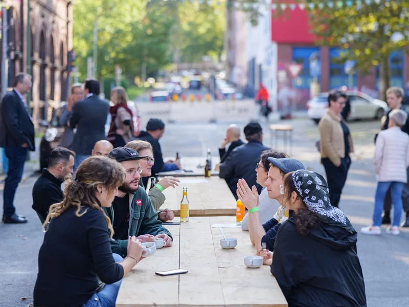 Menschen sitzen an Holzbänken vor der Quartiershalle der KoFabrik in Bochum.