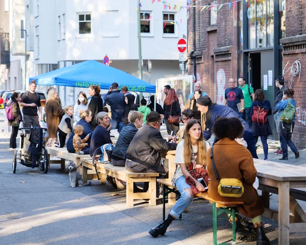 Menschen sitzen unter Sonnenschirmen zusammen auf Holzbänken vor der Quartiershalle und unterhalten sich fröhlich