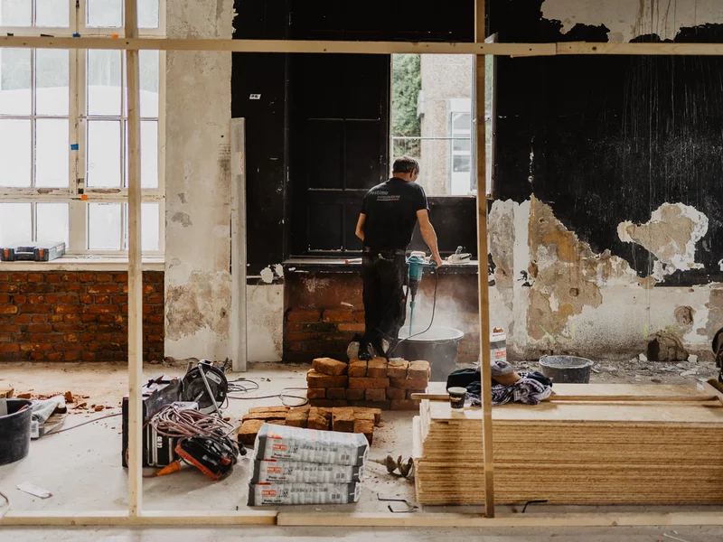 In einem Gebäude ist ein Holzrahmen im Hintergrund steht ein schwarz gekleideter Mensch, der mit einem Gerät in einem Eimer rührt. In der Bildmitte liegen Baumaterialien, Maschinen, drei Zementsäcke, Holzspanplatten, und aufgeschichtete Ziegelsteine