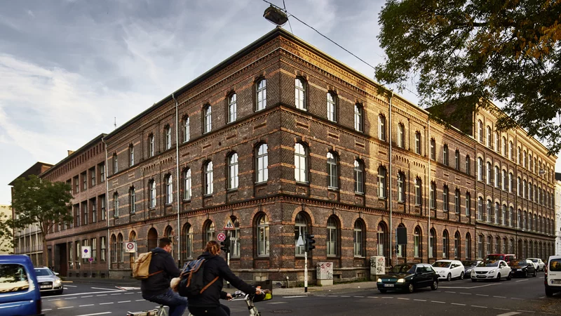 Ansicht der Samtweberei in Krefeld von der Ecke, ein Backsteingebäude. Im Vordergrund fahren zwei Menschen auf Fahrrädern vorbei, Autos parken auf der Straße.