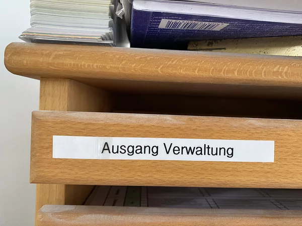 Ein Holzregal mit einem Aufkleber mit der Aufschrift Ausgang Verwaltung. Obenauf liegen Dokumentstapel und eine Zeitschrift.