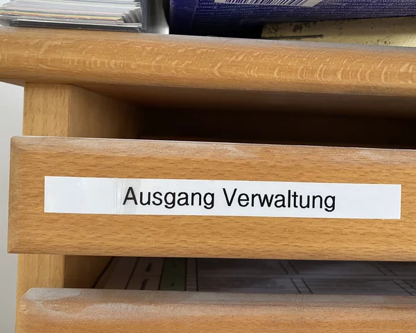 Ein Holzregal mit einem Aufkleber mit der Aufschrift Ausgang Verwaltung. Obenauf liegen Dokumentstapel und eine Zeitschrift.