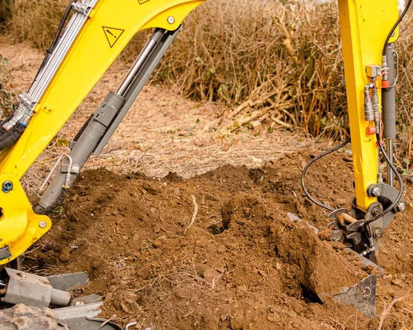Links im Bild steht ein kleiner gelber Bagger. Der Bagger gräbt einen lockeren Boden um.