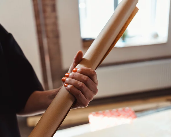 Eine Person mit orangenem Nagellack hält eine Rolle Packpapier. Im Hintergrund ist ein Fenster zu sehen.