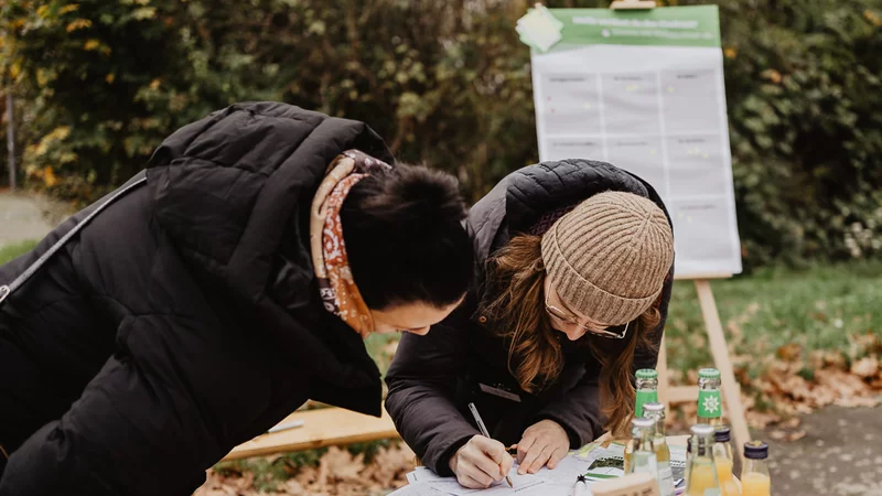 Zwei Personen beugen sich über einen Tisch, der draußen steht. Die rechts stehende Person trägt etwas auf einen Zettel ein, die links stehende Person schaut ihr dabei zu. Im Hintergrund steht eine Staffelei mit einer Information.