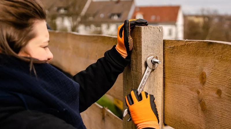 Eine Person mit orangenen Handschuhen befestigt einen Holzzaun mit einem Schraubenschlüssel. Im fernen HIntergrund sind einige Wohnhäuser zu sehen.