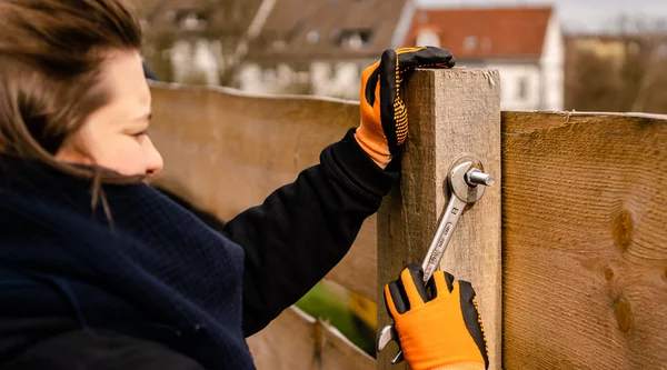 Eine Person mit orangenen Handschuhen befestigt einen Holzzaun mit einem Schraubenschlüssel. Im fernen HIntergrund sind einige Wohnhäuser zu sehen.