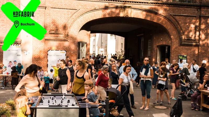 VIele Menschen vor einem Torbogen. Durch ein Logo und eine Ortsangabe ist das Bild deutlich als KoFabrik in Bochum gekennzeichnet.