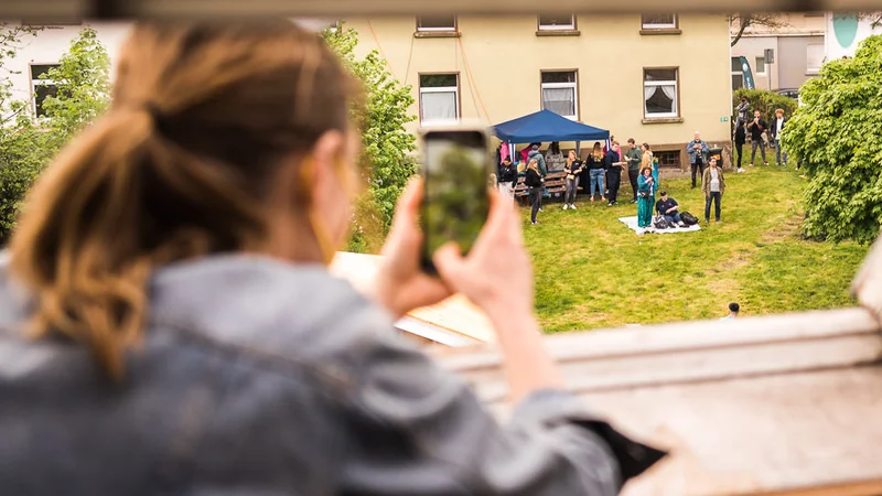Eine Person lehnt aus einem geöffneten Dachfenster und macht mit einem Handy ein Foto von einer Szene im Hintergrund. Hier sind einige Menschen um einen Pavillon versammelt.