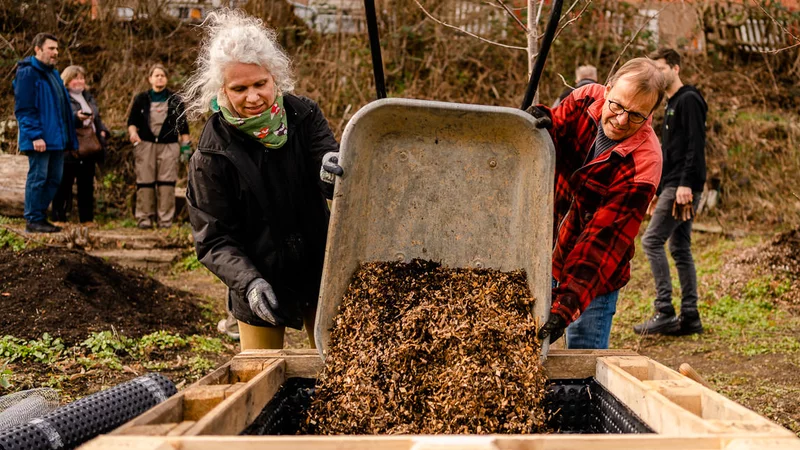Zwei Personen kippen gemeinsam Erde aus einer Schubkarre in ein Hochbeet aus Holzpaletten. Im Hintergrund stehen einige Personen im Garten. Ganz hinten ist eine kahle Hecke und eine leicht orangefarbene Häuserfront.