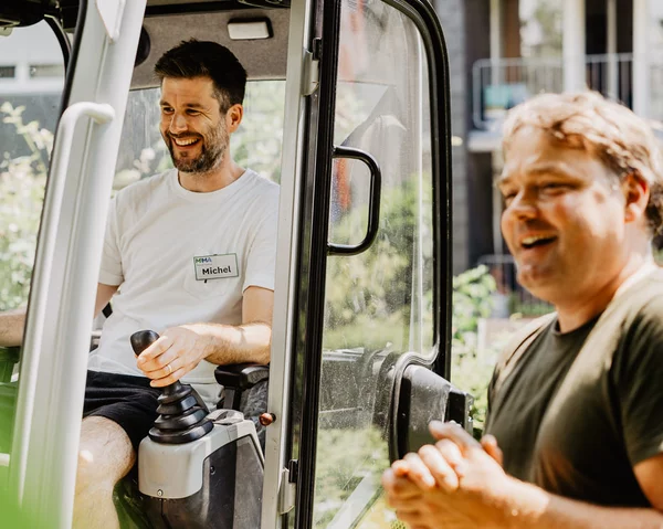 Mensch sitzt in Gartentraktor, fröhlicher Mensch steht daneben