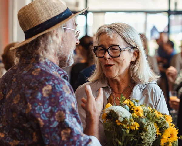 Drei Personen sind im Vordergrund in der Nachbarschaftsetage des BOB CAMPUS. Weiter hinten stehen mehr Menschen. Die Person vorne in der Mitte hält einen Blumenstrauß mit Sonnenblumen in der Hand. Die Person links von ihr trägt einen Strohhut. Sie unterhalten sich freudig.