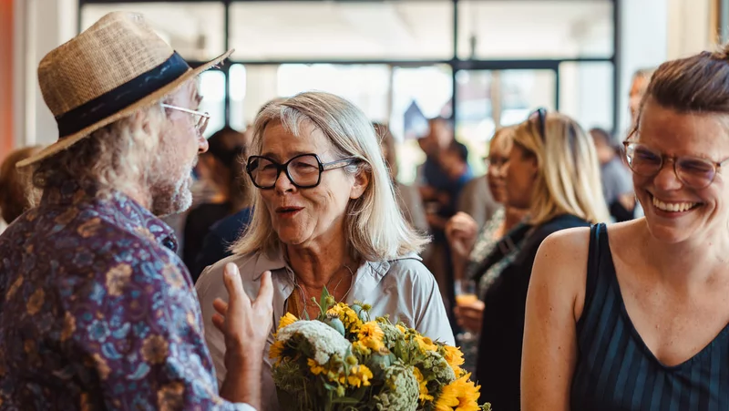 Drei Personen sind im Vordergrund in der Nachbarschaftsetage des BOB CAMPUS. Weiter hinten stehen mehr Menschen. Die Person vorne in der Mitte hält einen Blumenstrauß mit Sonnenblumen in der Hand. Die Person links von ihr trägt einen Strohhut. Sie unterhalten sich freudig.