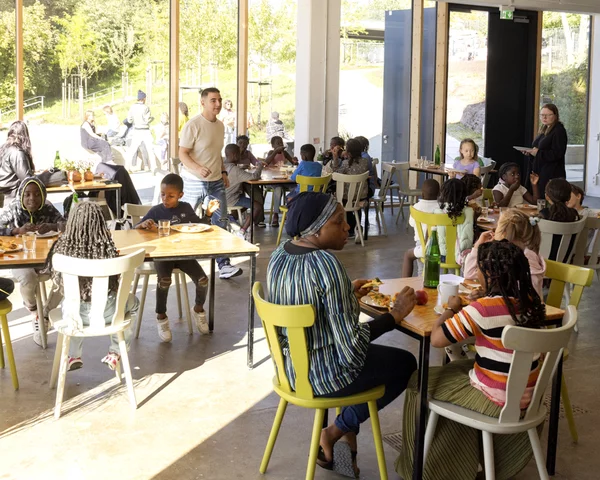 In einem Gastraum sitzen in Tischgruppen auf bunten Holzstühlen mehrere Menschen zusammen und essen, einige von ihnen sind Kinder. Im Hintergrund sieht man durch eine Glasfront in den grünen Nachbarschaftspark Oberbarmen.