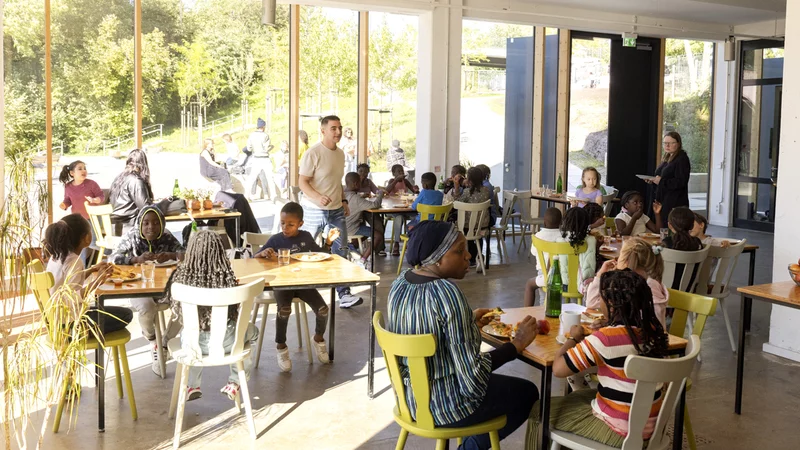In einem Gastraum sitzen in Tischgruppen auf bunten Holzstühlen mehrere Menschen zusammen und essen, einige von ihnen sind Kinder. Im Hintergrund sieht man durch eine Glasfront in den grünen Nachbarschaftspark Oberbarmen.