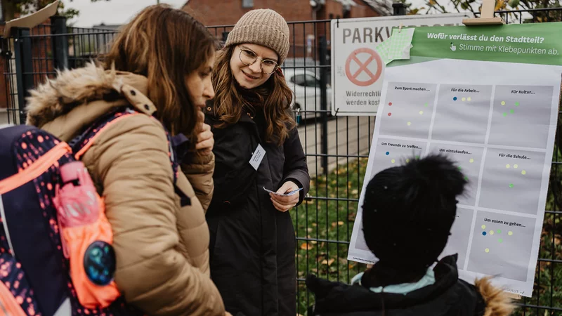 Drei Personen stehen draußen vor einer Tafel mit Informationen zum Einfüllen. Die Person links hat einen Schulranzen auf dem Rücken, rechts steht ein Kind mit schwarzer Pudelmütze.