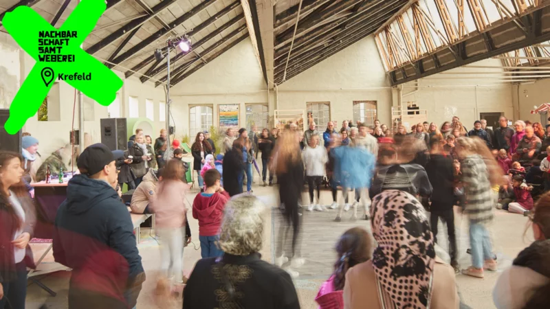 Viele Menschen in der Shedhalle. Durch ein Logo und eine Ortsangabe ist das Bild deutlich als Nachbarschaft Samtweberei in Krefeld gekennzeichnet.