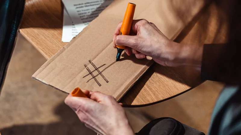 Eine Person schreibt das Symbol Hashtag mit schwarzem Edding auf ein Stück Pappkarton. Als Unterlage dient ein Holzstuhl.