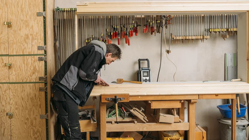 Eine Werkbank mit darüber aufgehängtem Werkzeug ist zu sehen. Links daneben steht ein Schrank mit Holzspantüren und am linken Bildrand lehnen Holzleisten. Neben der Werkbank links, etwa in der Bildmitte schaut ein Handwerker auf seine Arbeit.