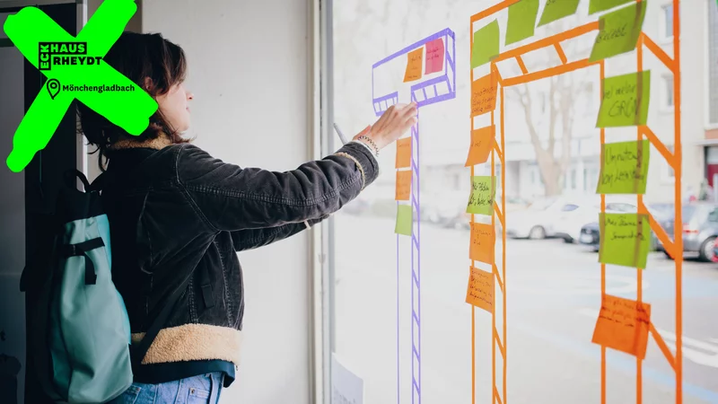 Eine Person in der linken Bildhälfte steht mit ihrer linken Seite zur Kamera. Die Person klebt mit ihrer rechten Hand ein Post-it von innen an ein Schaufenster. Auf dem Schaufenster kleben bereits mehrere bunte Post-its.