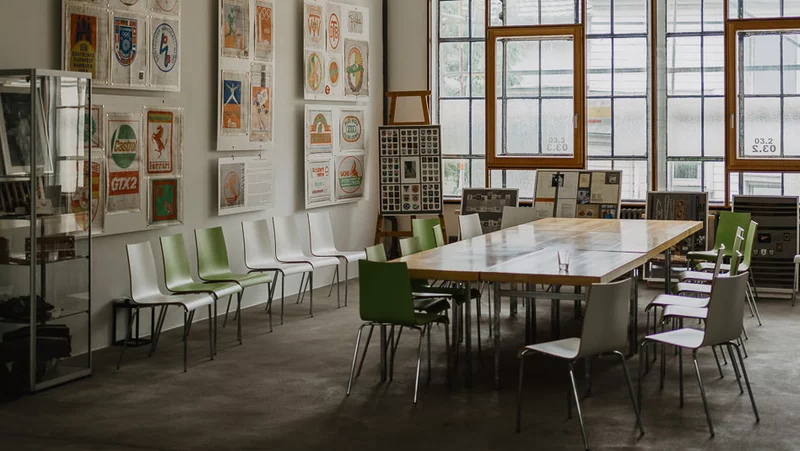 Ein Innenraum mit einem Besprechungstisch und etwa fünfzehn Stühlen. An der Wand links hängen bunte Poster. HInter dem Tisch sind zwei große Fenster.