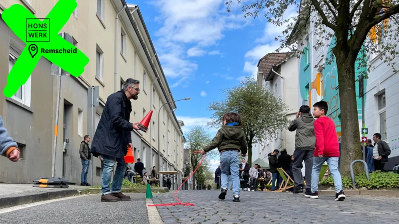 Kinder spielen auf der Siemensstraße, im Hintergrund sitzen Erwachsene an Tischen. Durch ein Logo und eine Ortsangabe ist das Bild deutlich als HONSWERK in Remscheid gekennzeichnet.