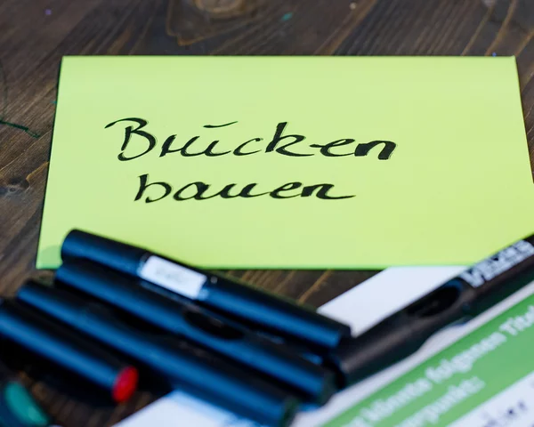 Auf einem Tisch liegen Haftnotizen und Marker. Im Zentrum liegt ein grüner Haftnotiz-Block mit der schwarzen Aufschrift Brücken bauen.