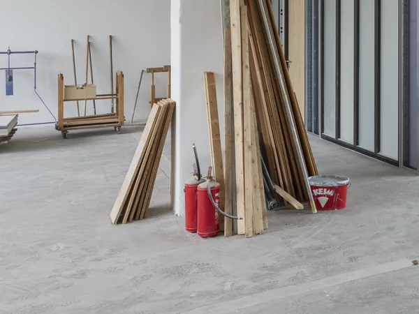 In einem Innenraum ist ein Raum im Rohbau eingebaut. An der rechten Seite sind große Außenfenster. An einer Säule mitten im Raum lehnen Holzleisten, ein Farbeimer und zwei Feuerlöscher. Rechts steht ein abmontierter Heizkörper. 