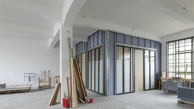 In einem Innenraum ist ein Raum im Rohbau eingebaut. An der rechten Seite sind große Außenfenster. An einer Säule mitten im Raum lehnen Holzleisten, ein Farbeimer und zwei Feuerlöscher. Rechts steht ein abmontierter Heizkörper. 