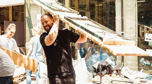 Fröhliche Menschen auf einer Baustelle tragen Gartentische/Bierbänke