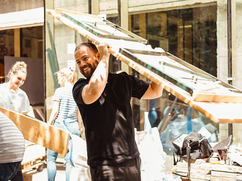Fröhliche Menschen auf einer Baustelle tragen Gartentische/Bierbänke