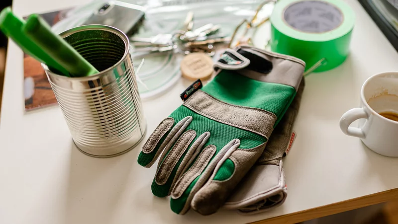 Auf einem Tisch steht eine KOnservendose in der eine Zange mit grünem Griff steckt. Außerdem grüne Gartenhandschuhe, ein grünes Tape und eine Tasse Kaffee.