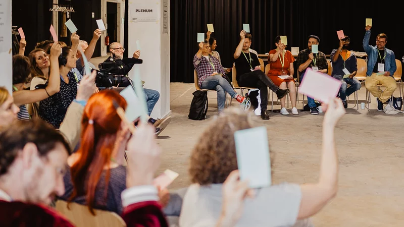 Menschen halten in einem großen Versammlungsraum in einem Stuhlkreis Stimmkarten hoch