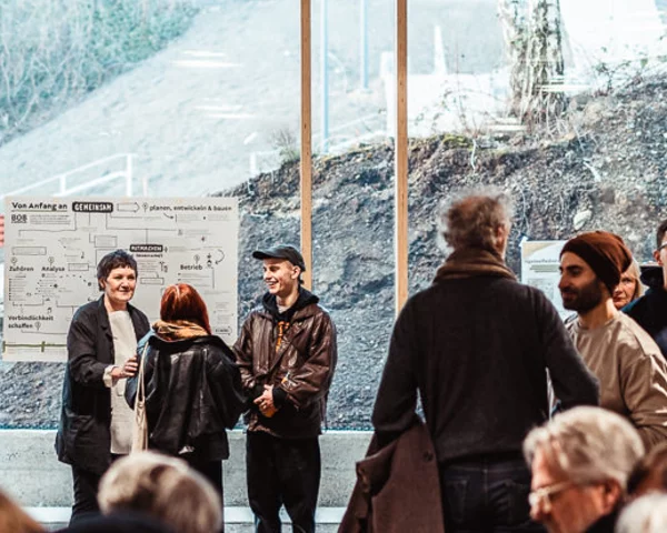 Eine große Gruppe Menschen steht in der Nachbarschaftsetage des BOB CAMPUS in Wuppertal Oberbarmen. Im Hintergrund eine Fensterfront, durch die der Nachbarschaftspark sichtbar ist.