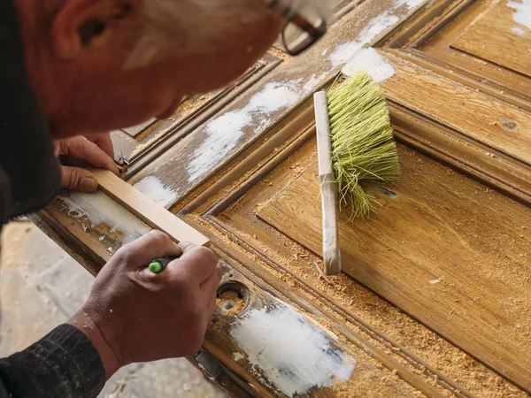 Ein Handwerker beugt sich über eine alte Holztür, auf der weiße Farbereste zu sehen sind. Auf der Tür liegt ein Handfeger mit hellgrünen Borsten und einige Holzspäne. Der Arbeiter zeichnet etwas an.