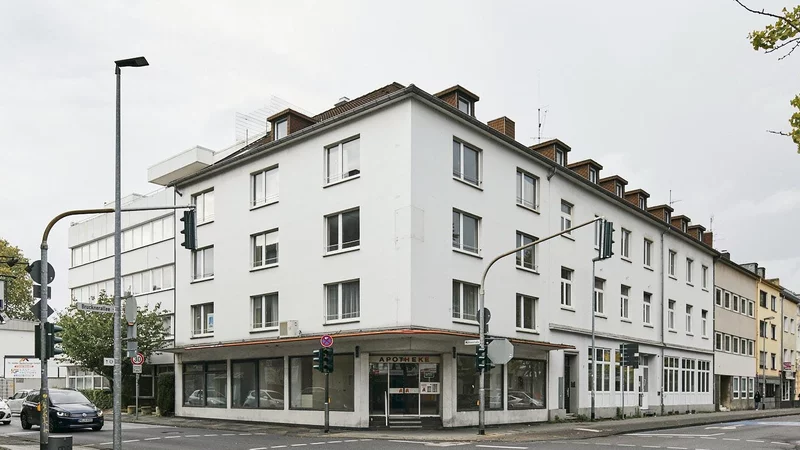 Das Eckhaus Rheydt von vorne fotografiert an einer Straßenecke gelegen. Die Erdgeschosszone ist klar erkennbar. Darüber drei Geschosse mit symmetrischen Fenstern und Giebelfenstern. Links ist nach hinten versetzt die ehemalige Wirtschaftsschule an der Mühlenstraße erkennbar.