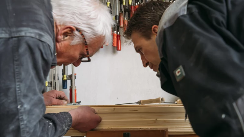 Zwei Arbeiter beugen sich über eine Detail Holzarbeit. Im Hintergrund hängt Werkzeug.
