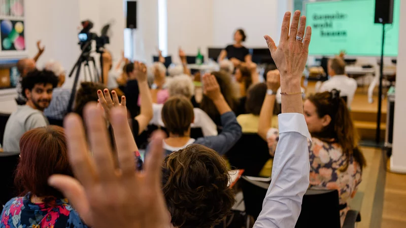 Viele Menschen sitzen in Stuhlreihen, einige heben ihre Hände zur Abstimmung. Im Hintergrund ist auf einer Leinwand undeutlich auf grünem Hintergrund in schwarz Gemeinwohl bauen zu lesen.