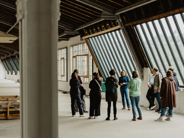 EIne Gruppe von Menschen steht unter einer hohen Holzdecke in einer ehemaligen Fabrikhalle. Rechts ist eine große schräge Fensterfront. Im Vordergrund des Bildes eine trahende Säule. 