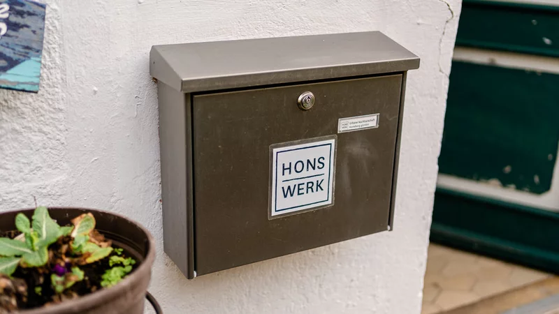 Links ein Blumenkasten, in der Mitte ein silberner Briefkasten mit dem Logo des HONSWERK an einer weißen Hauswand, rechts daneben steht eine grüne Tür offen.