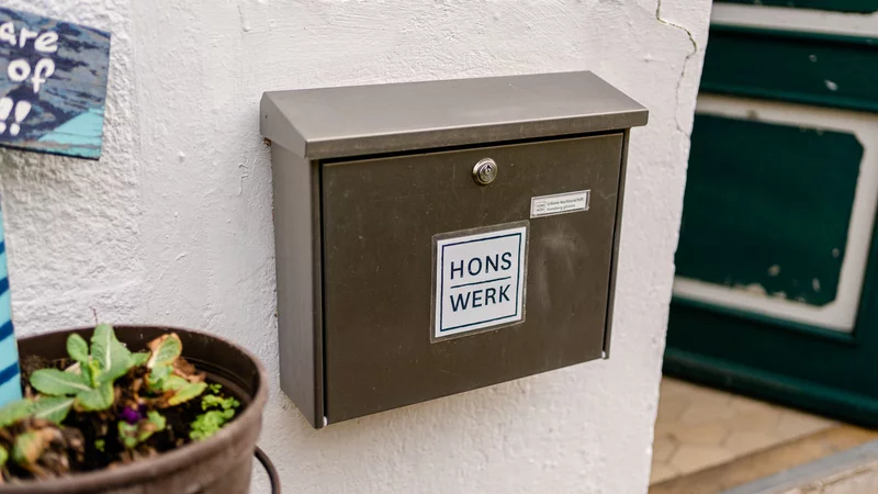 Links ein Blumenkasten, in der Mitte ein silberner Briefkasten mit dem Logo des HONSWERK an einer weißen Hauswand, rechts daneben steht eine grüne Tür offen.