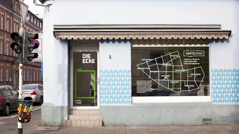 Ein Ladenlokal an einer Hausecke mit einer Glastür, zu der einige Stufen hinauf führen,  und einem großen Glasfenster. Tür und Fenster sind großflächig beschrieben.